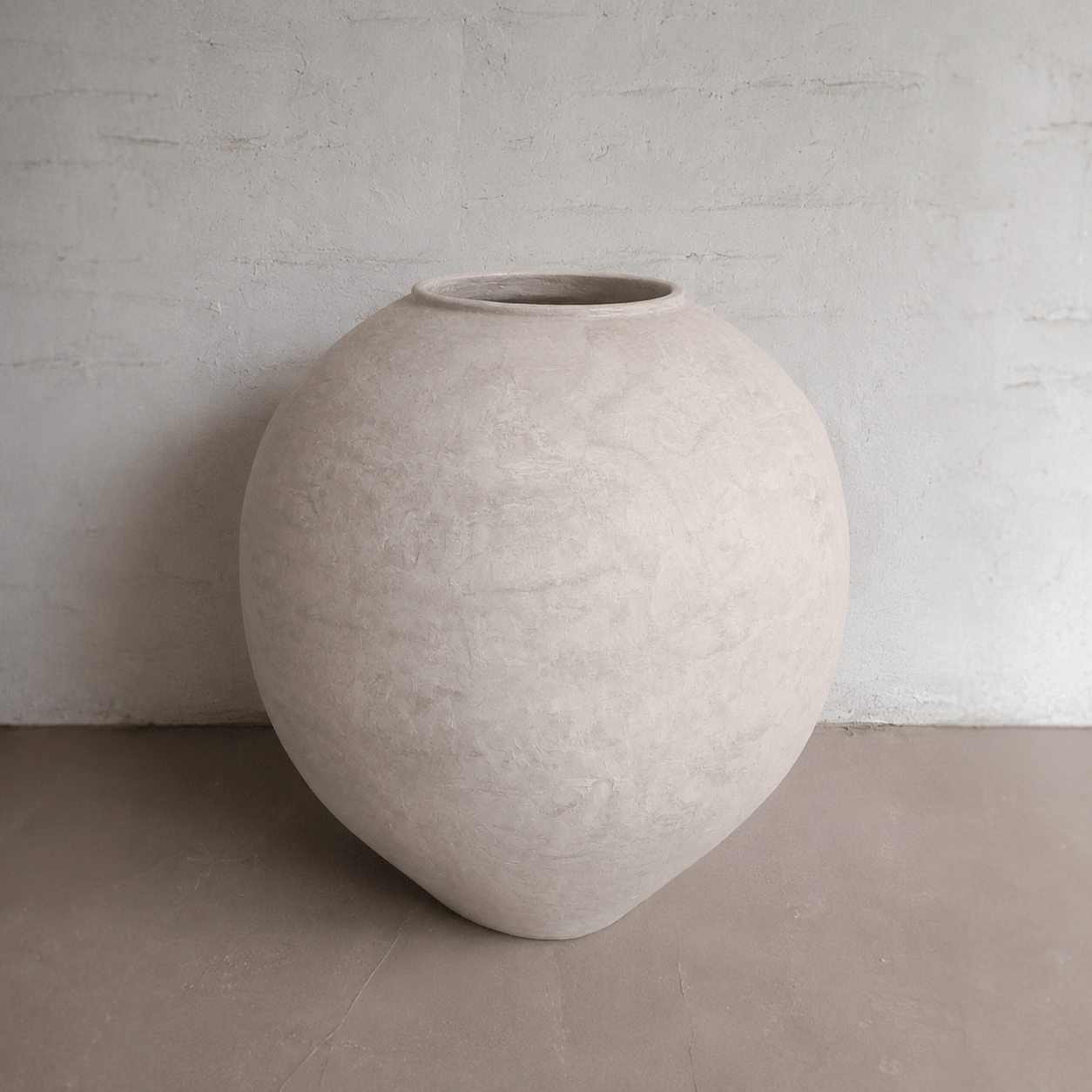 The Syracuse Organic Fiberglass Pot - Earth Range by Florastyle By Hingham, a large round light beige pot with a narrow opening, rests on a smooth floor against a textured off-white wall.