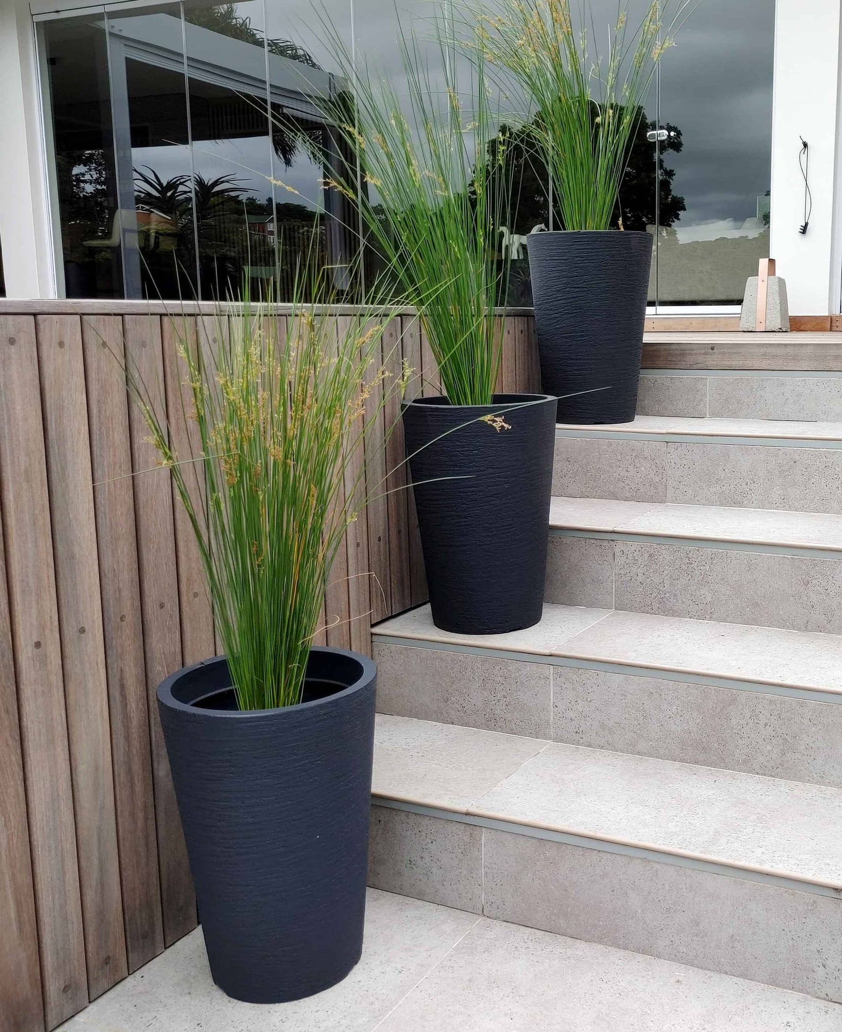European Conic Japi plant pots. Displayed on a flight of stairs, outdoors, the small in the picture adding interest in an outdoor space. Planted up with Aristida junciformis. These pots are slim and come in three different heights. Colour options are available. Shown here is the colour lead. Florastyle by Hingham. This style fits in with any style of decor.