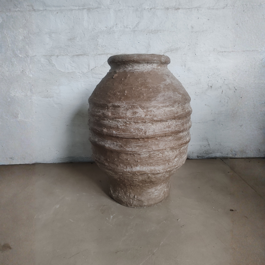The Sparta Organic Fiberglass Pot - Earth Range by Florastyle By Hingham, featuring a wide body, narrow neck, and horizontal ridges, sits on a plain floor against a white textured wall.