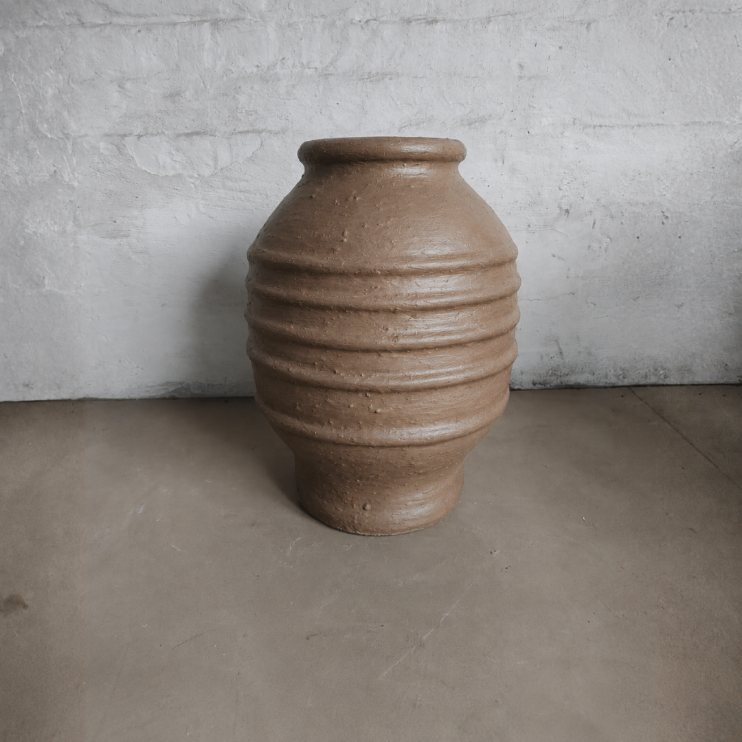 A large Sparta Organic Fiberglass Pot from Florastyle By Hingham, in an earth-tone finish with horizontal ridges, sits on a gray concrete floor against a white, rough brick wall.