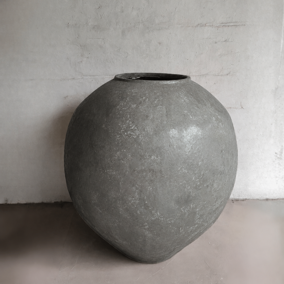 The Syracuse Organic Fiberglass Pot – Earth Range by Florastyle By Hingham is a large, round gray planter with a weathered texture, wide body, and narrow opening. It sits against a plain, light wall on a smooth floor.