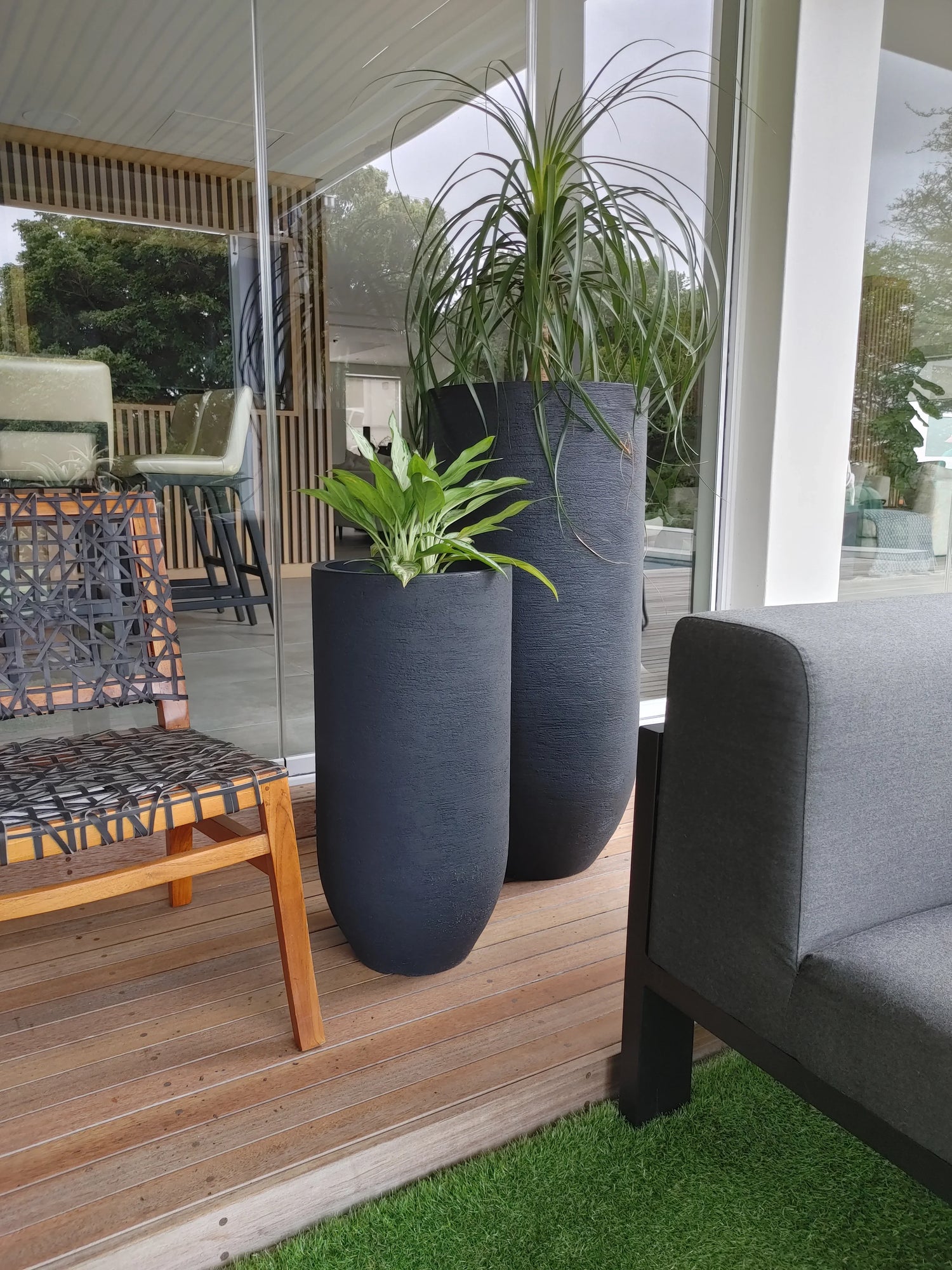 Two tall black planters with green plants sit on a wooden deck next to a gray outdoor sofa and a wooden chair, in front of large glass windows reflecting trees and greenery.