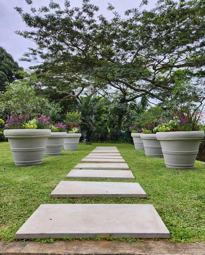 Pathway made of concrete slabs leading through a garden with large Japi flower pots and trees.