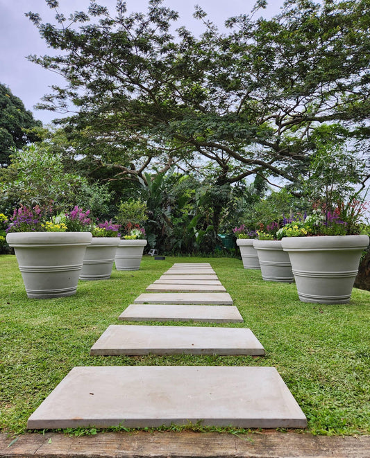 Pathway made of concrete slabs leading through a garden with large Japi flower pots and trees.