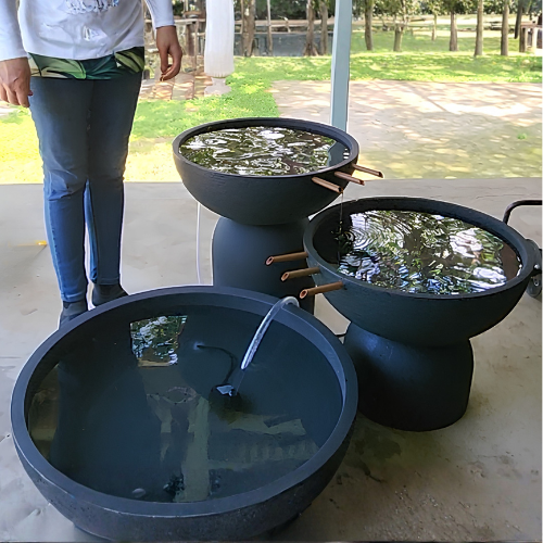 A person stands by the Florastyle By Hingham Water Feature - 3 Tier European Bowl with Large Pond, featuring round black polyresin basins—two elevated on stands with water pipes and one on the ground—outdoors on a covered patio.