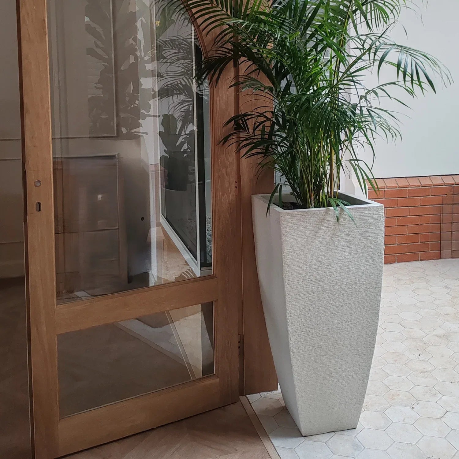 Tall square, textured, Off White, tapering shaped Japi planter.  Placed by a door frame. Palms planted inside. 