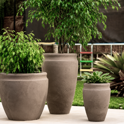 Three large Florastyle By Hingham Antique Japi Planters, including an urn-style design, display leafy green plants on a patio with a lush garden and blurred background behind them.