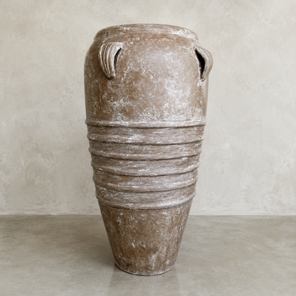 The Athens Organic Fiberglass Pot - Earth Range by Florastyle By Hingham is a large, brown, weathered pot with two small top handles and horizontal mid ridges, standing on a smooth floor against a light textured wall.