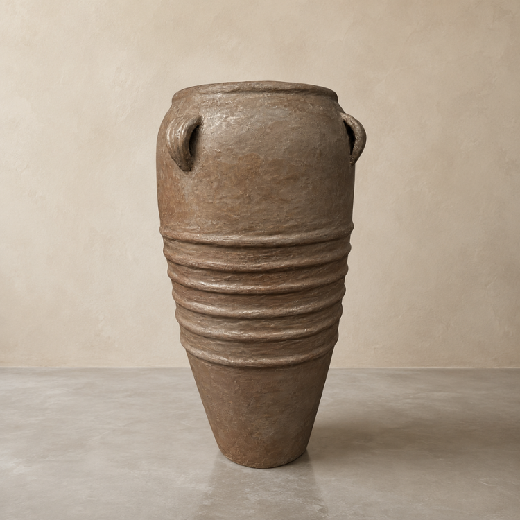 The Athens Organic Fiberglass Pot – Earth Range by Florastyle By Hingham features ancient Greek-inspired design with a conical base, textured lower ridges, and two loop handles, shown here on a plain floor against a beige wall.