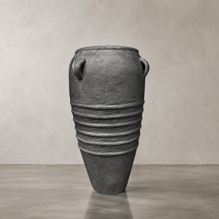 The Athens Organic Fiberglass Pot from Florastyle By Hingham features a tall, dark gray finish with ribbed bands and two small handles near the rim, displayed on a smooth floor against a light textured wall.