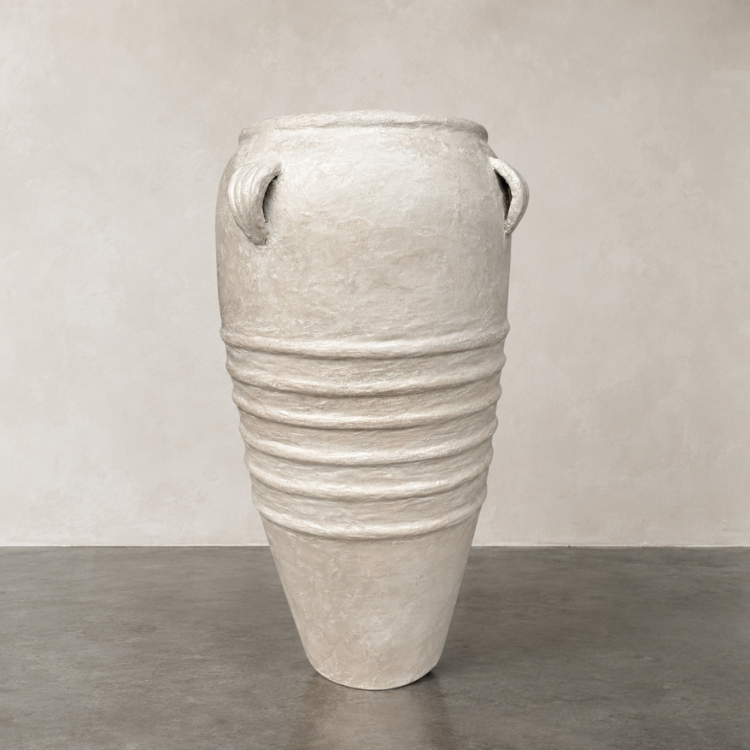 The Athens Organic Fiberglass Pot - Earth Range by Florastyle By Hingham features a tall, textured design with dual handles and horizontal ridges, shown on a gray concrete floor against a light beige wall.