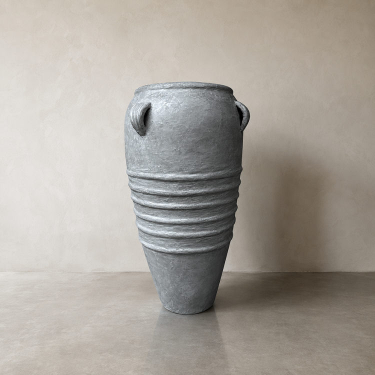 The Athens Organic Fiberglass Pot from Florastyle By Hingham, part of the Earth Range, features a tall gray finish and side handles reminiscent of ancient Greek pottery, displayed against a beige wall on a concrete floor.