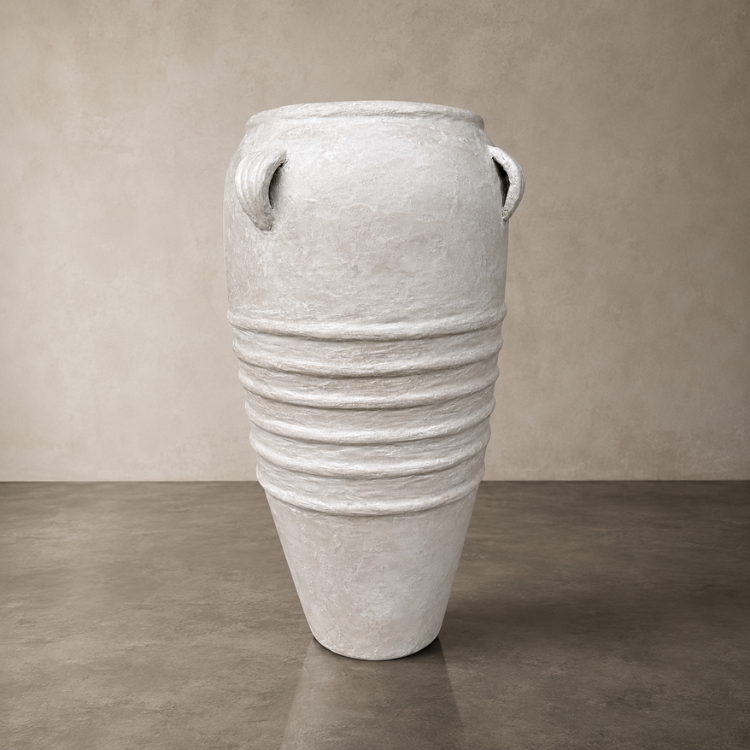 The Athens Organic Fiberglass Pot - Earth Range by Florastyle By Hingham stands on a plain concrete floor against a neutral beige wall, featuring horizontal ridges, a tapered base, and small handles near the rim for an ancient Greek-inspired look.