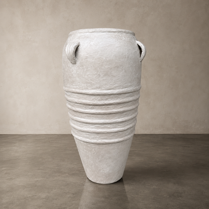 The Athens Organic Fiberglass Pot - Earth Range by Florastyle By Hingham stands on a plain concrete floor against a neutral beige wall, featuring horizontal ridges, a tapered base, and small handles near the rim for an ancient Greek-inspired look.
