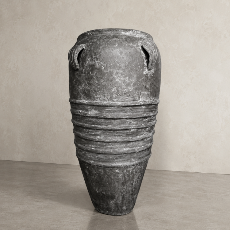 The Athens Organic Fiberglass Pot - Earth Range by Florastyle By Hingham, a tall gray planter with a narrow base, wide middle, and two small handles near the rim, stands on a smooth floor against a beige textured wall.