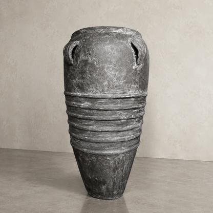 The Athens Organic Fiberglass Pot - Earth Range by Florastyle By Hingham, a tall gray planter with a narrow base, wide middle, and two small handles near the rim, stands on a smooth floor against a beige textured wall.