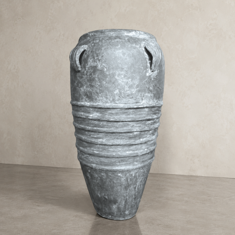 The Athens Organic Fiberglass Pot - Earth Range by Florastyle By Hingham features a tall, weathered gray design with horizontal ridges and two small loop handles, evoking ancient Greek pottery, displayed on a smooth floor against a beige wall.