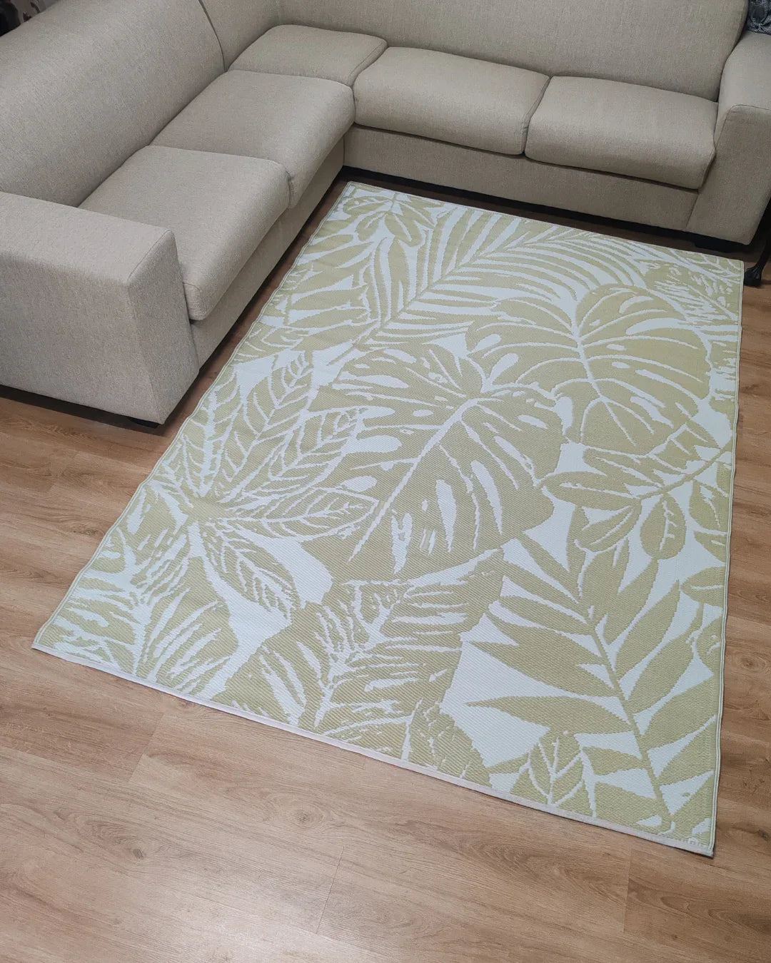 A Large Bali Outdoor Reversible Mat by Florastyle By Hingham, lightweight and water-resistant with a beige and white leafy design, is laid on a light wood floor between two parts of a light-colored L-shaped sofa.