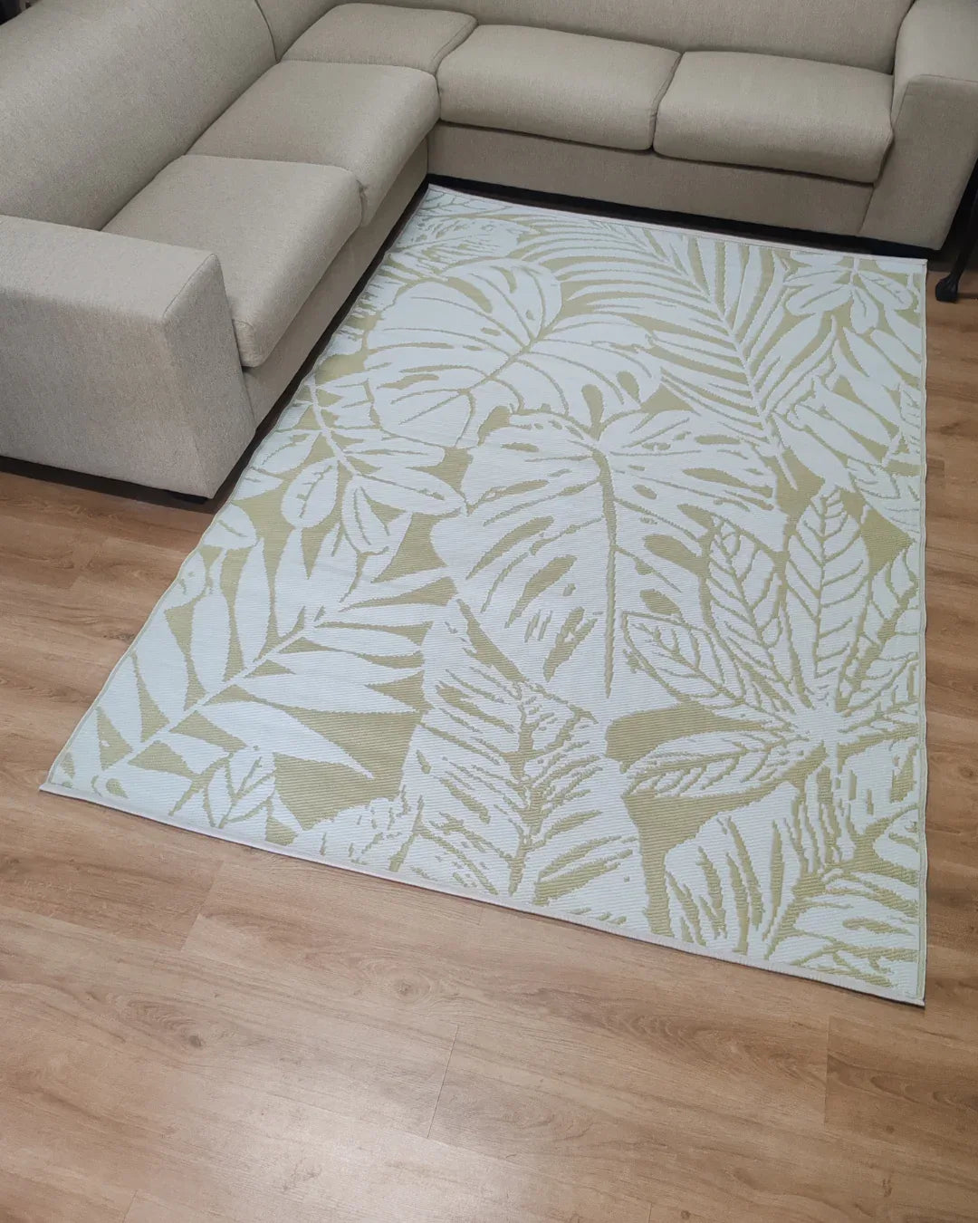 A lightweight beige sectional sofa surrounds the Florastyle By Hingham Large Bali Outdoor Reversible Mat, featuring a pale green and white tropical leaf pattern, placed on light wood flooring.