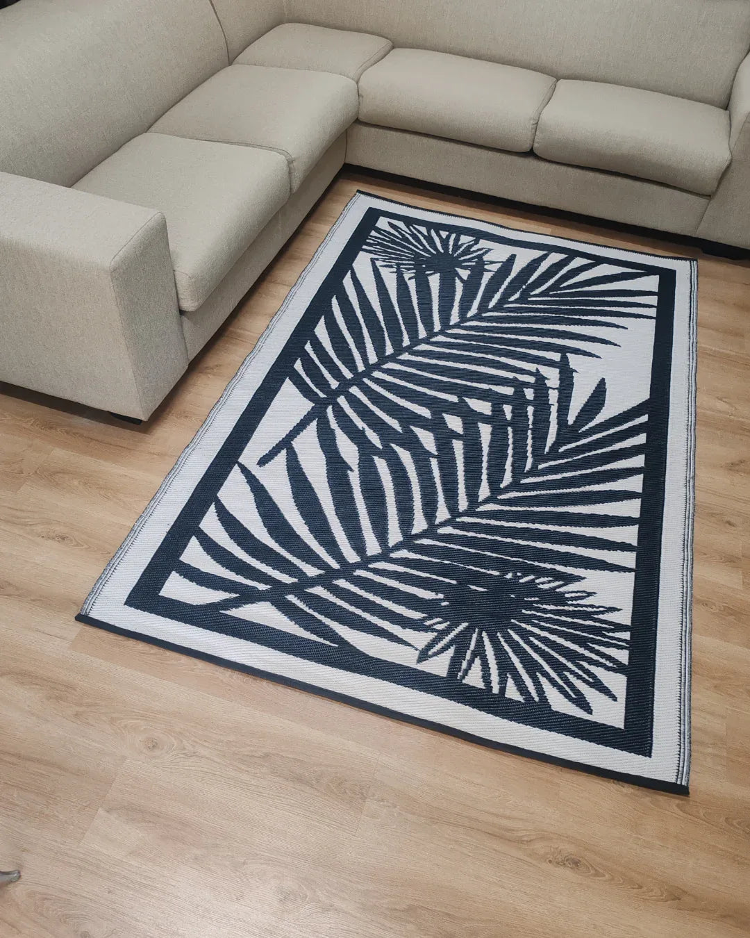 A lightweight beige sectional sofa surrounds the Florastyle By Hingham Medium Cairo Outdoor Reversible Mat, featuring a black and white palm leaf pattern, placed on a light wood floor.