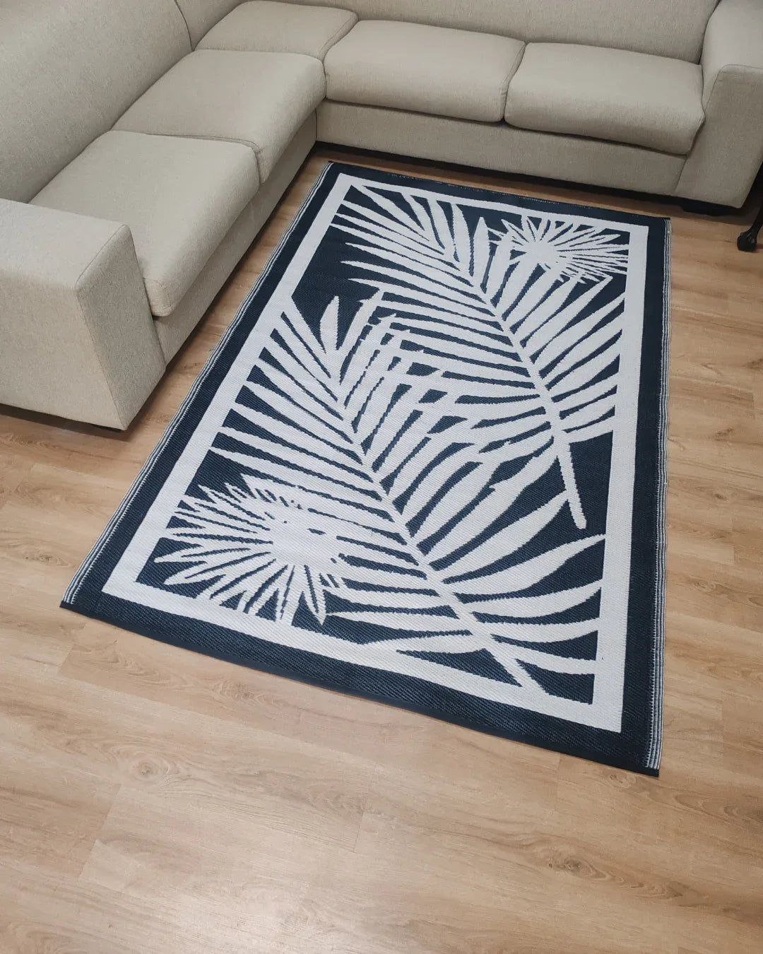 A Medium Cairo Outdoor Reversible Mat by Florastyle By Hingham, featuring a lightweight double-woven design and water resistance, with a black background and white tropical leaf pattern, is placed on a light wooden floor between beige sectional sofas.