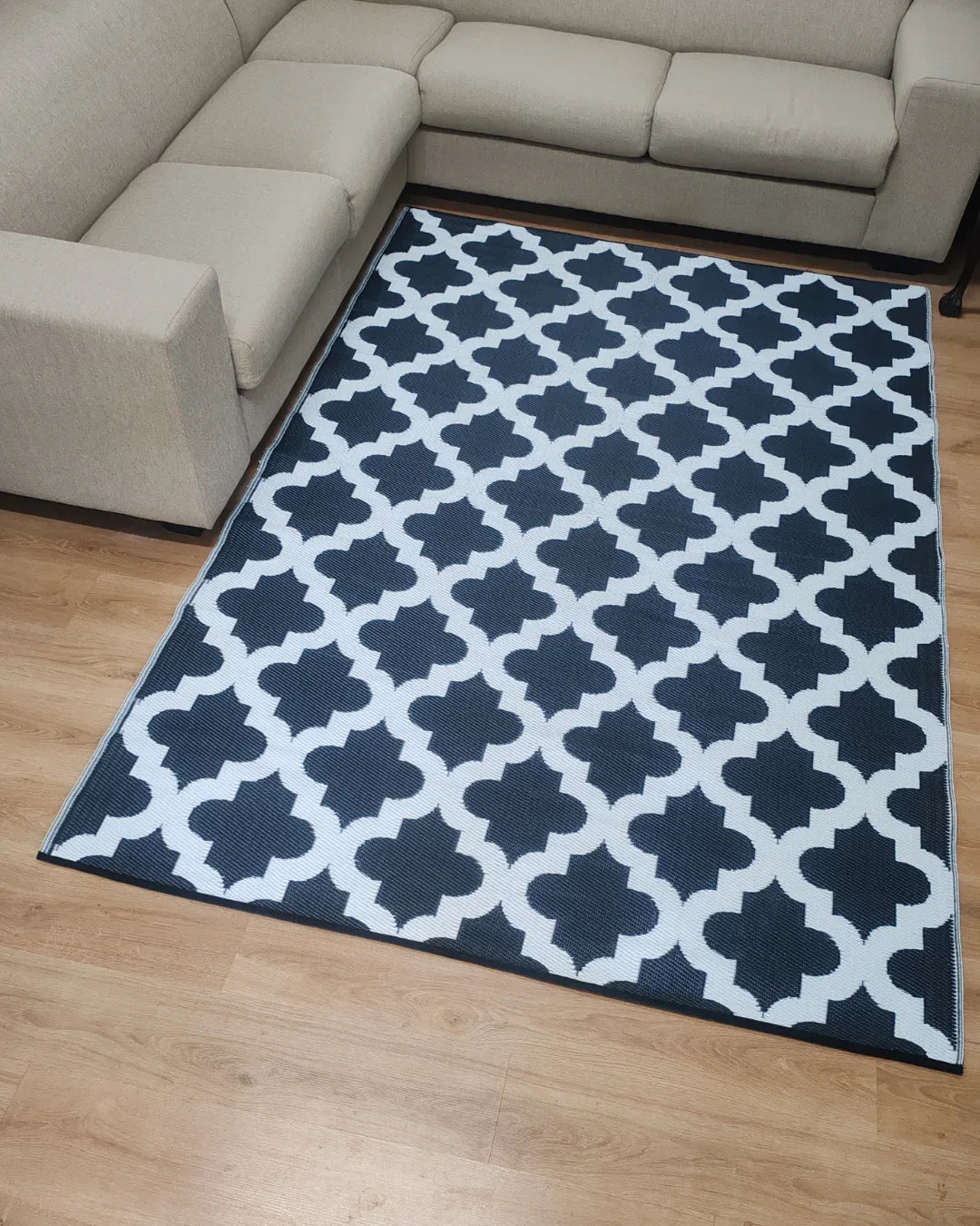 A lightweight beige sectional sofa surrounds the Large Casablanca Outdoor Reversible Mat by Florastyle By Hingham, featuring a black and white Moroccan geometric pattern, placed on a light wood floor.