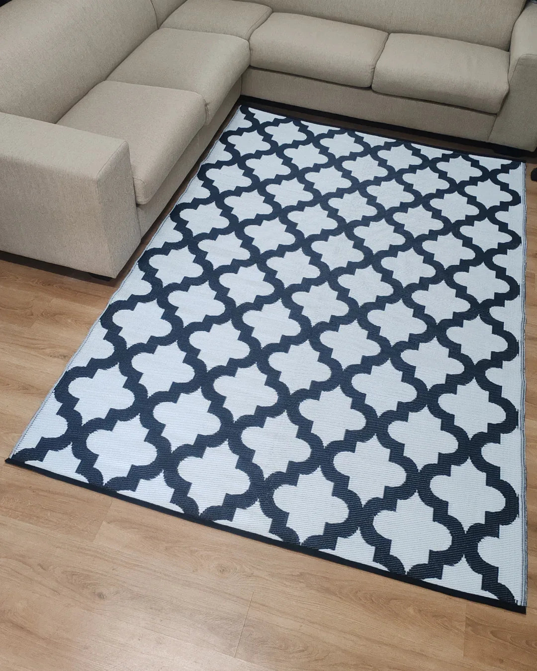 A beige, lightweight sectional sofa surrounds the Florastyle By Hingham Large Casablanca Outdoor Reversible Mat—a double-woven, water-resistant area rug with a white background and black geometric trellis pattern—on a light wood floor.