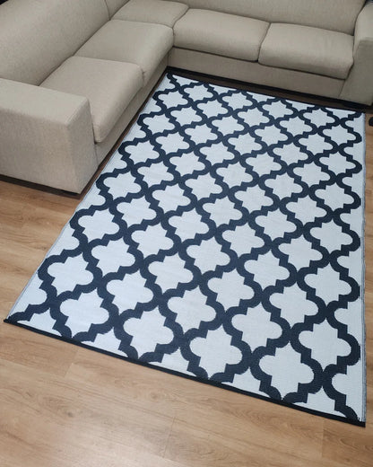 A beige, lightweight sectional sofa surrounds the Florastyle By Hingham Large Casablanca Outdoor Reversible Mat—a double-woven, water-resistant area rug with a white background and black geometric trellis pattern—on a light wood floor.