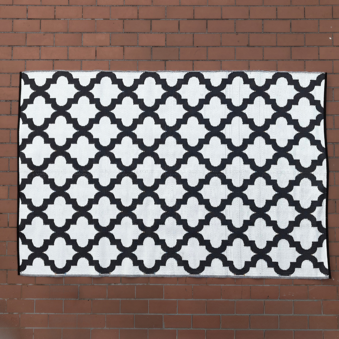 The Large Casablanca Outdoor Reversible Mat by Florastyle By Hingham, a lightweight double-woven rug with a black and white geometric lattice pattern, is laid flat on a red brick surface.