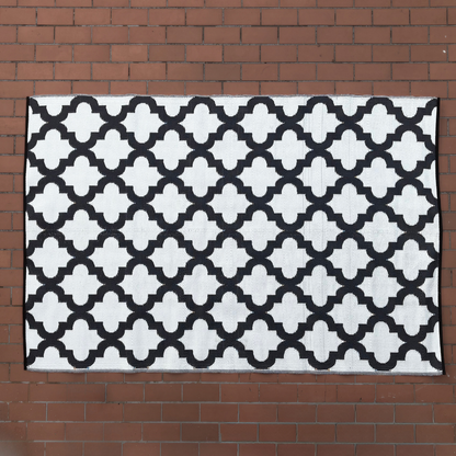 The Large Casablanca Outdoor Reversible Mat by Florastyle By Hingham, a lightweight double-woven rug with a black and white geometric lattice pattern, is laid flat on a red brick surface.