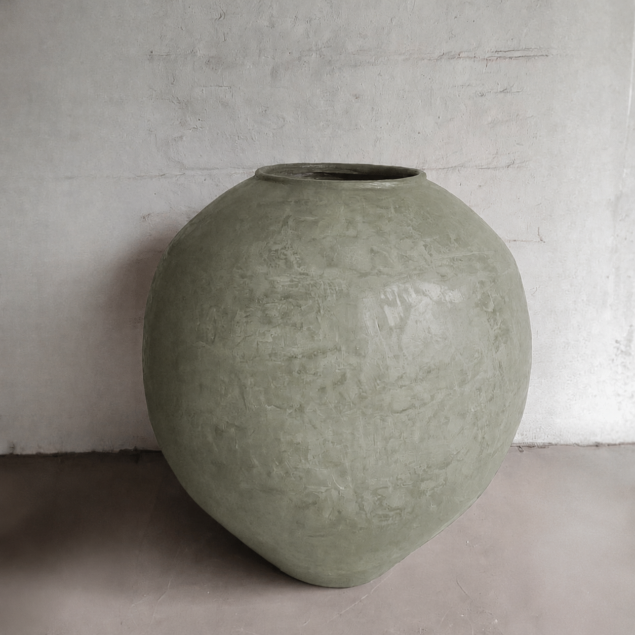 The Syracuse Organic Fiberglass Pot - Earth Range by Florastyle By Hingham, large and round in matte olive-green with a rough finish, sits on a smooth floor against a lightly textured white wall.