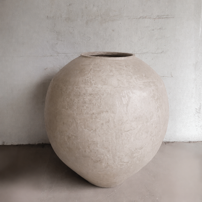 The Syracuse Organic Fiberglass Pot from Florastyle By Hingham's Earth Range, featuring a large, round design and rough-textured surface, stands on a smooth floor against a plain light-colored wall.
