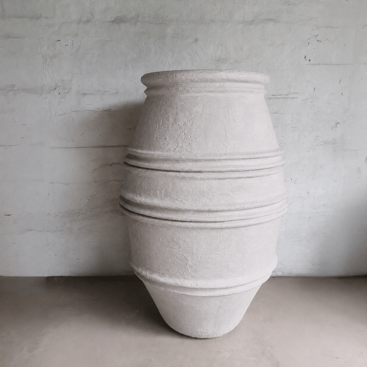 The Crete Organic Fiberglass Pot - Earth Range by Florastyle By Hingham features a large, textured cylindrical design with central ridged bands, standing upright against a light gray wall—ideal for organic modern decor.