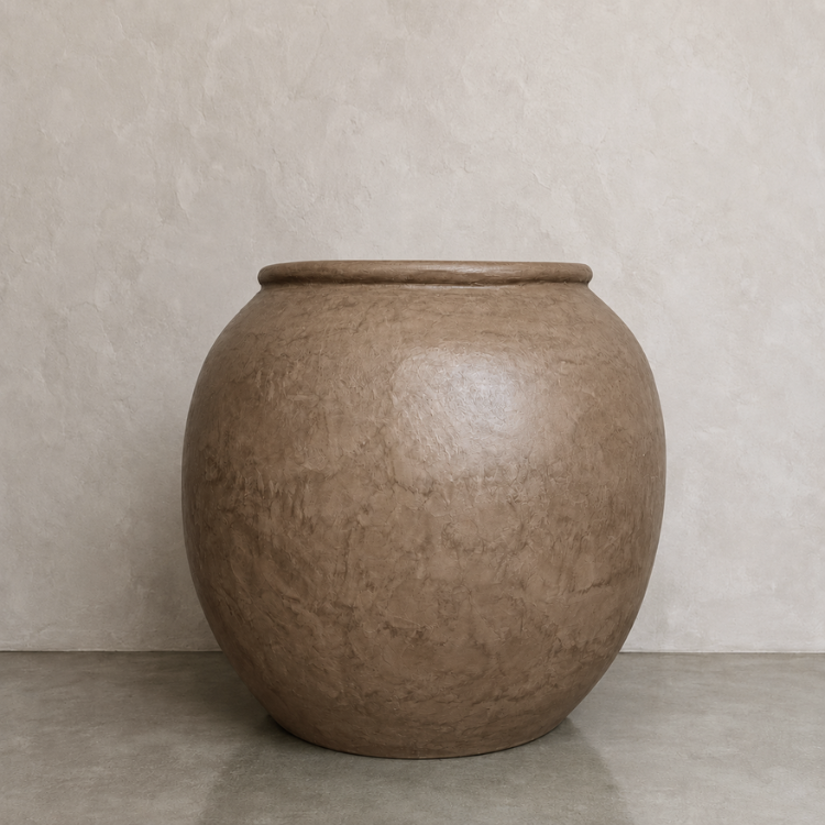 A large, round Delphi Organic Fiberglass Pot from the Earth Range by Florastyle By Hingham, with a wide opening and earthy brown finish, sits on a smooth gray surface against a textured beige wall, perfectly evoking the timeless appeal of an indoor plant pot.