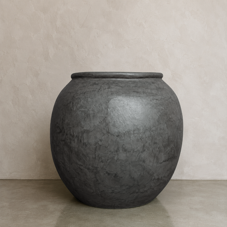 A large, round, dark gray Delphi Organic Fiberglass Pot from the Earth Range by Florastyle By Hingham, featuring a wide opening, sits on a smooth surface against a light beige textured wall.