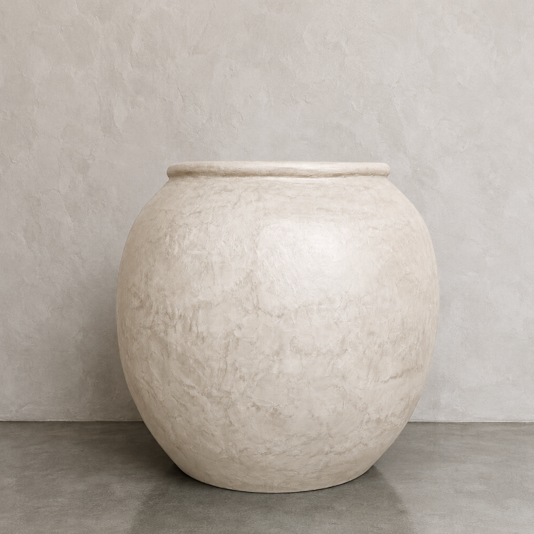 A large, round, beige Delphi Organic Fiberglass Pot from the Earth Range by Florastyle By Hingham sits on a gray concrete floor against a matching beige textured wall.