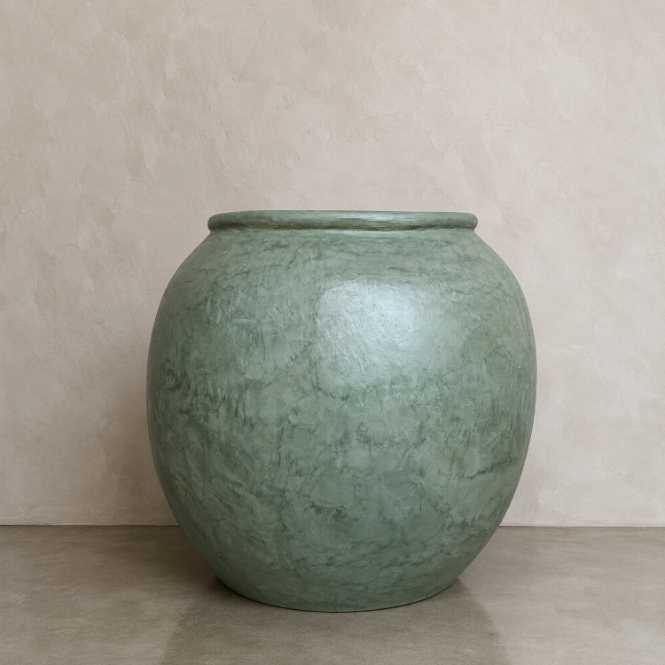 A large, round Delphi Organic Fiberglass Pot from Florastyle By Hingham’s Earth Range, featuring a smooth, glossy Vogue Green finish, sits on a beige floor against a textured light beige wall.