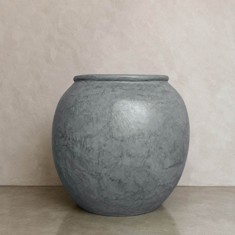 The Delphi Organic Fiberglass Pot - Earth Range by Florastyle By Hingham—a large, round, gray fiberglass planter with a slightly narrow opening—rests on a smooth surface against a light beige textured wall.