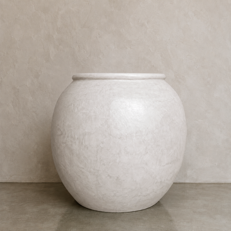 A large round white Delphi Organic Fiberglass Pot from Florastyle By Hingham’s Earth Range sits on a gray floor against a light beige textured wall.