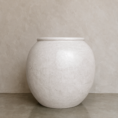 A large round white Delphi Organic Fiberglass Pot from Florastyle By Hingham’s Earth Range sits on a gray floor against a light beige textured wall.