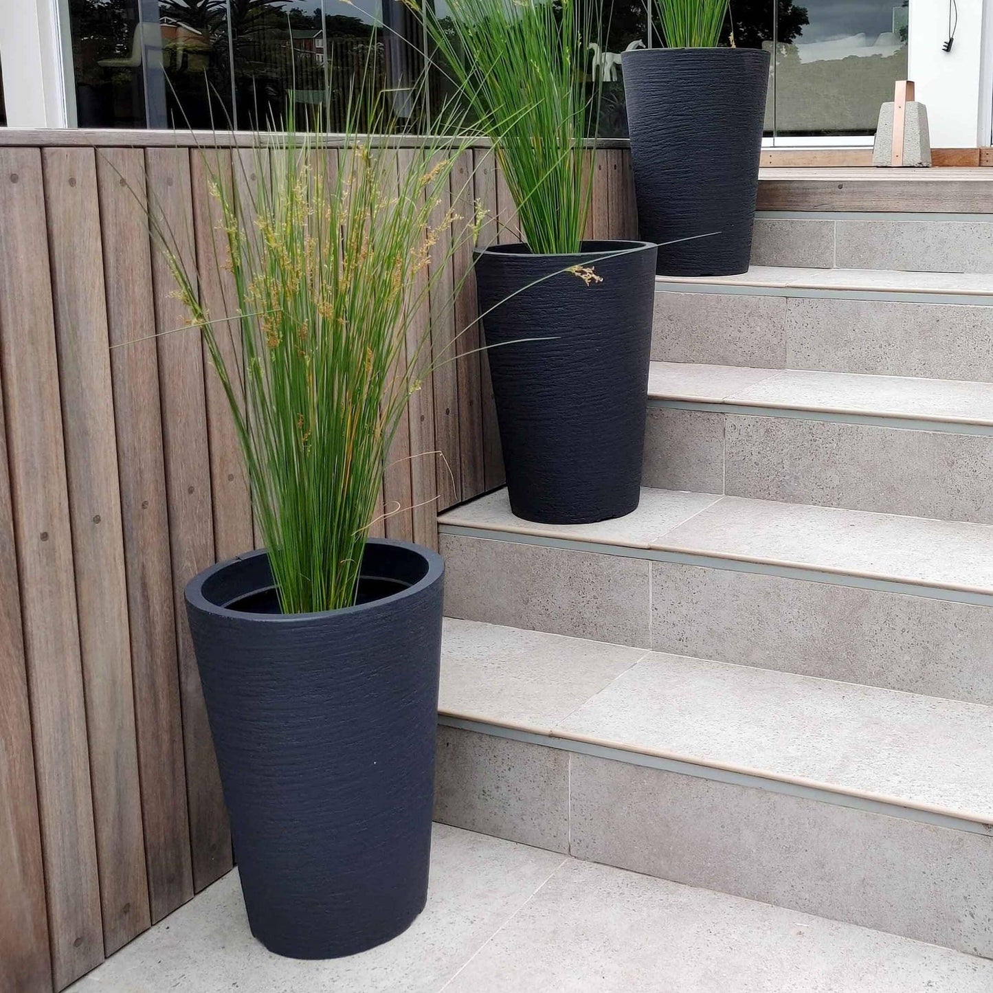 European Conic Japi plant pots. Displayed on a flight of stairs, outdoors, the small in the picture adding interest in an outdoor space. Planted up with Aristida junciformis. These pots are slim and come in three different heights. Colour options are available. Shown here is the colour lead. Florastyle by Hingham. This style fits in with any style of decor.