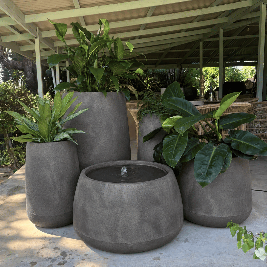 The Full Bios Set with One Bios Low Water Feature by Florastyle By Hingham creates a modern patio oasis, featuring lush plants and a sleek water feature beneath a covered structure with garden greenery as the backdrop.