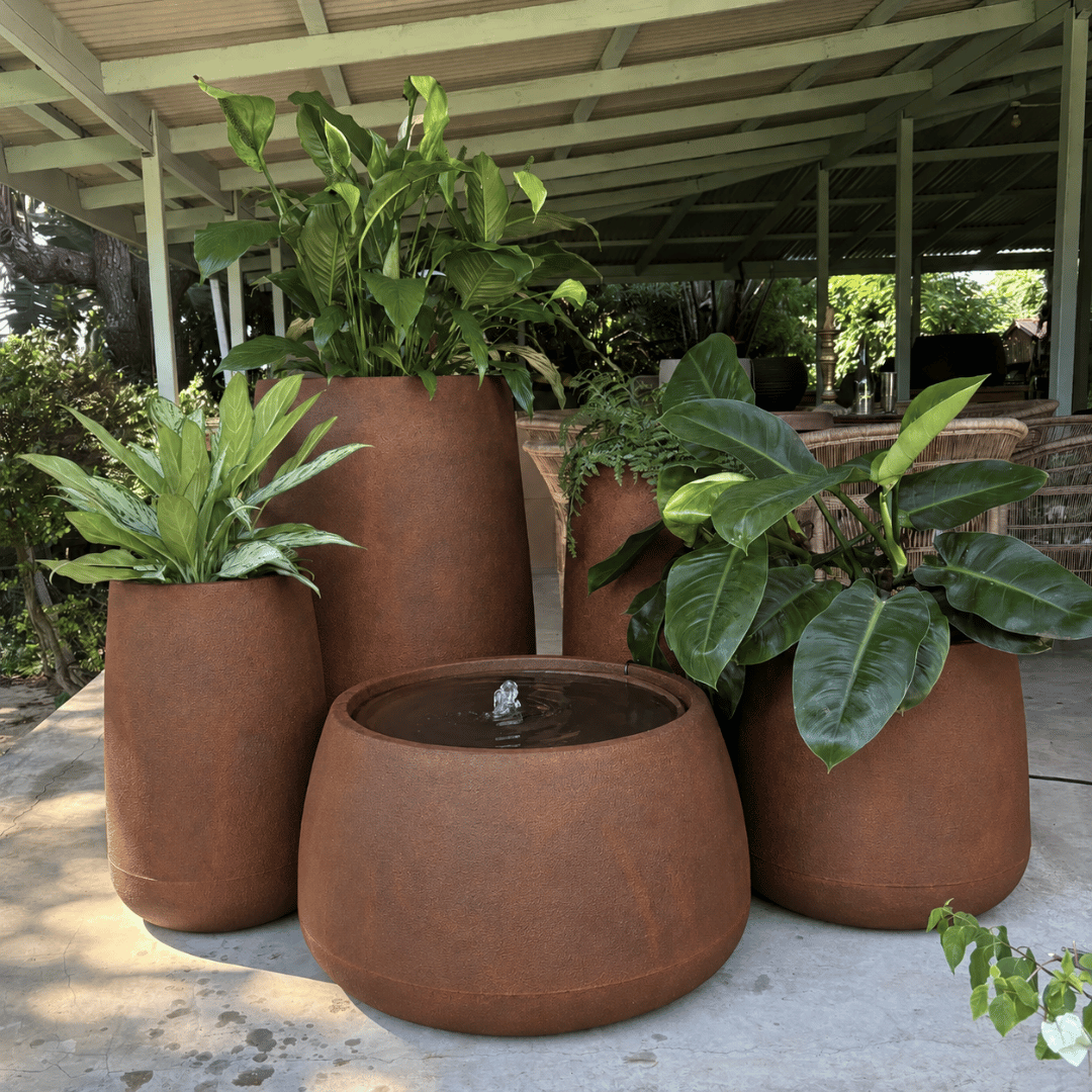 The Full Bios Set with One Bios Low Water Feature by Florastyle By Hingham features five large rust-colored planters and a small water feature, creating a modern oasis on a covered patio with lush plants and outdoor seating.