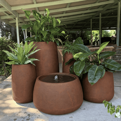 The Full Bios Set with One Bios Low Water Feature by Florastyle By Hingham features five large rust-colored planters and a small water feature, creating a modern oasis on a covered patio with lush plants and outdoor seating.