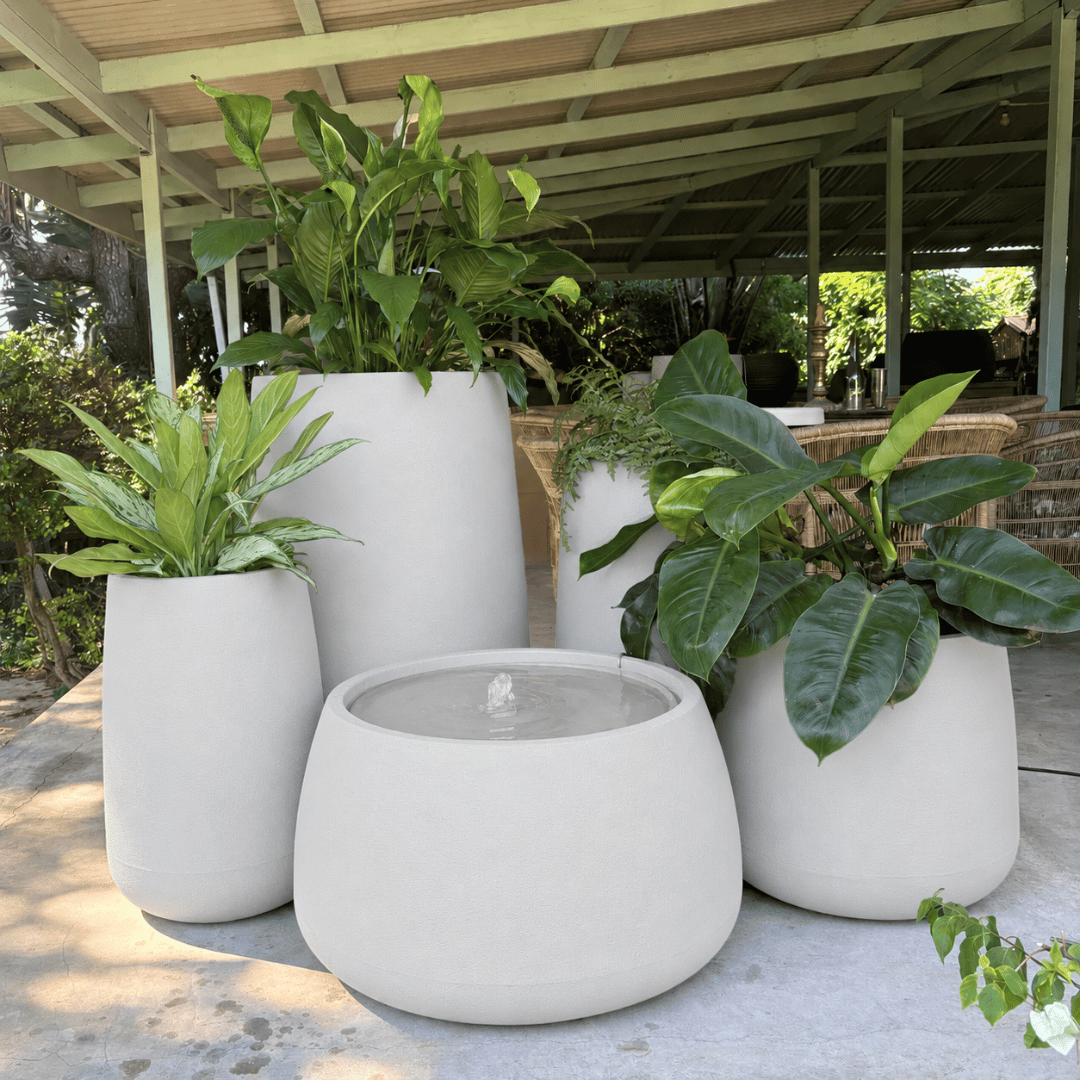 A Full Bios Set with One Bios Low Water Feature by Florastyle By Hingham—five large, modern, UV-resistant planters with assorted green plants—adorns a patio under a covered outdoor structure amid lush greenery.
