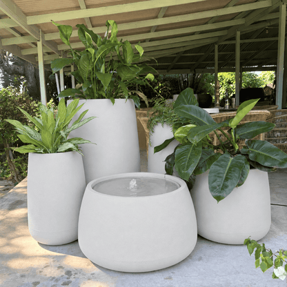 A Full Bios Set with One Bios Low Water Feature by Florastyle By Hingham—five large, modern, UV-resistant planters with assorted green plants—adorns a patio under a covered outdoor structure amid lush greenery.