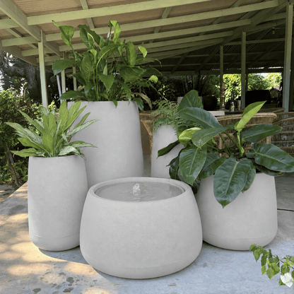 The Full Bios Set with One Bios Low Water Feature by Florastyle By Hingham, featuring five large modern planters and lush green plants, sits on a patio beneath a covered roof in a garden setting.