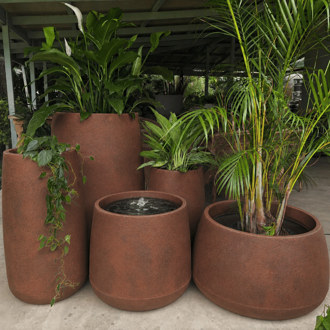 Large reddish-brown modern planters from Florastyle By Hingham’s Full Bios Set with One Bios Medium Water Feature, some holding lush tropical plants, are grouped together under a sheltered outdoor area.