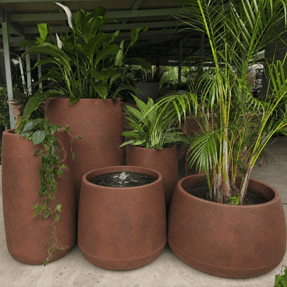 Large reddish-brown modern planters from Florastyle By Hingham’s Full Bios Set with One Bios Medium Water Feature, some holding lush tropical plants, are grouped together under a sheltered outdoor area.
