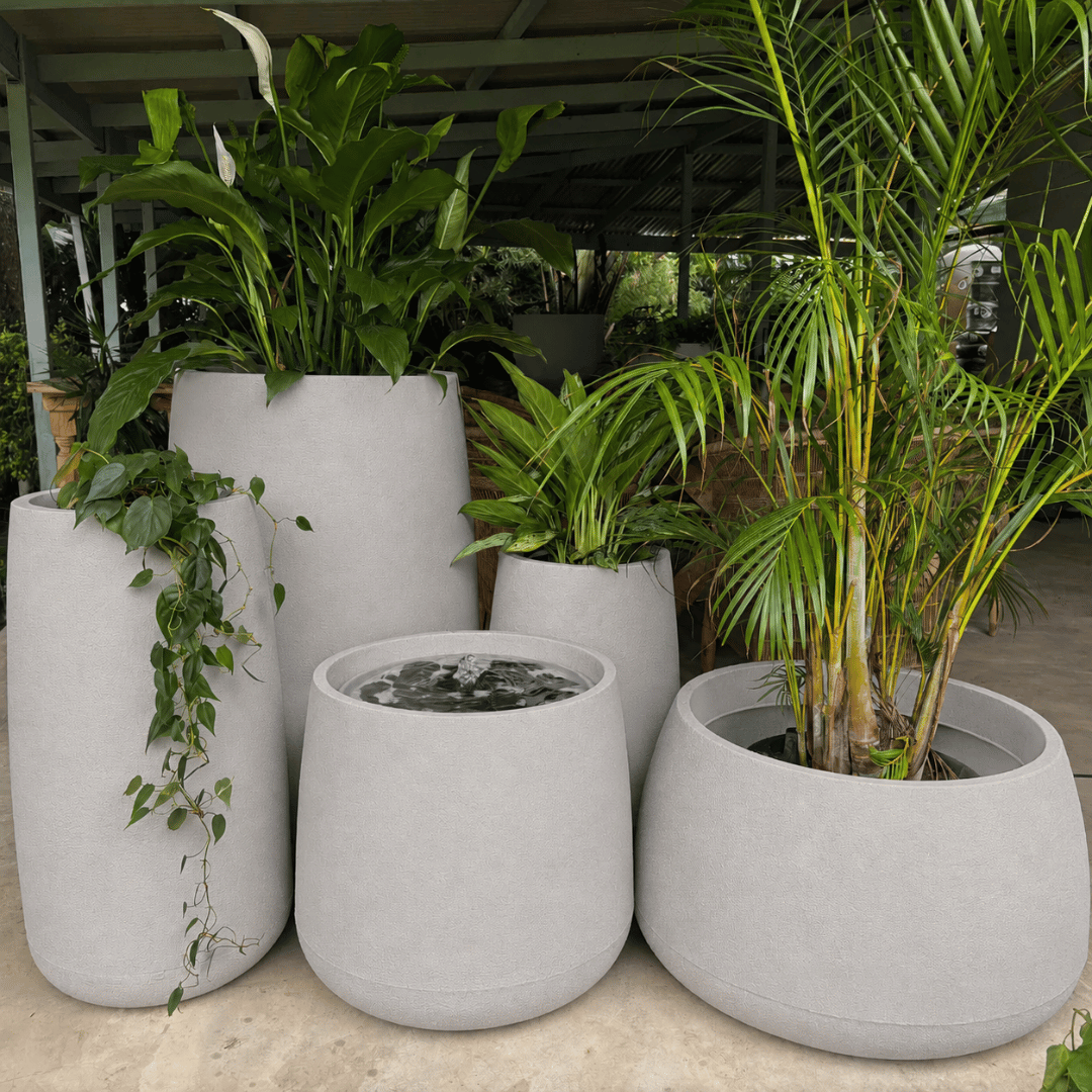The Florastyle By Hingham Full Bios Set with One Bios Medium Water Feature includes five large UV-resistant polyresin planters with lush plants, arranged on a concrete floor under a covered outdoor space.
